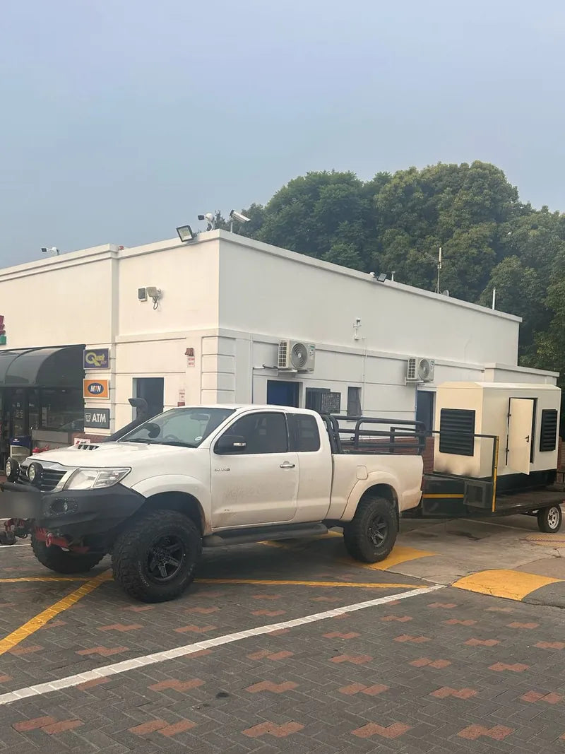 A white bakkie towing a generator on a trailer outside a commercial building