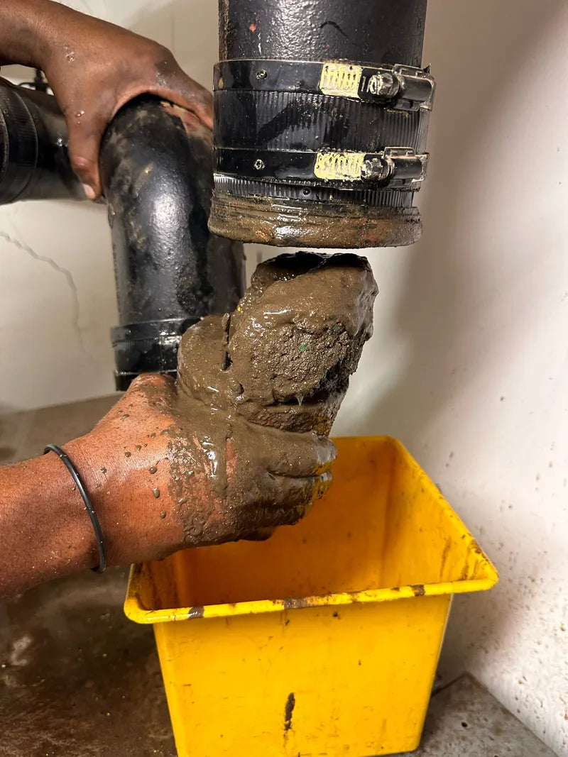 Hands pulling a plug of sludge out of a cast-iron waste pipe into a yellow bucket