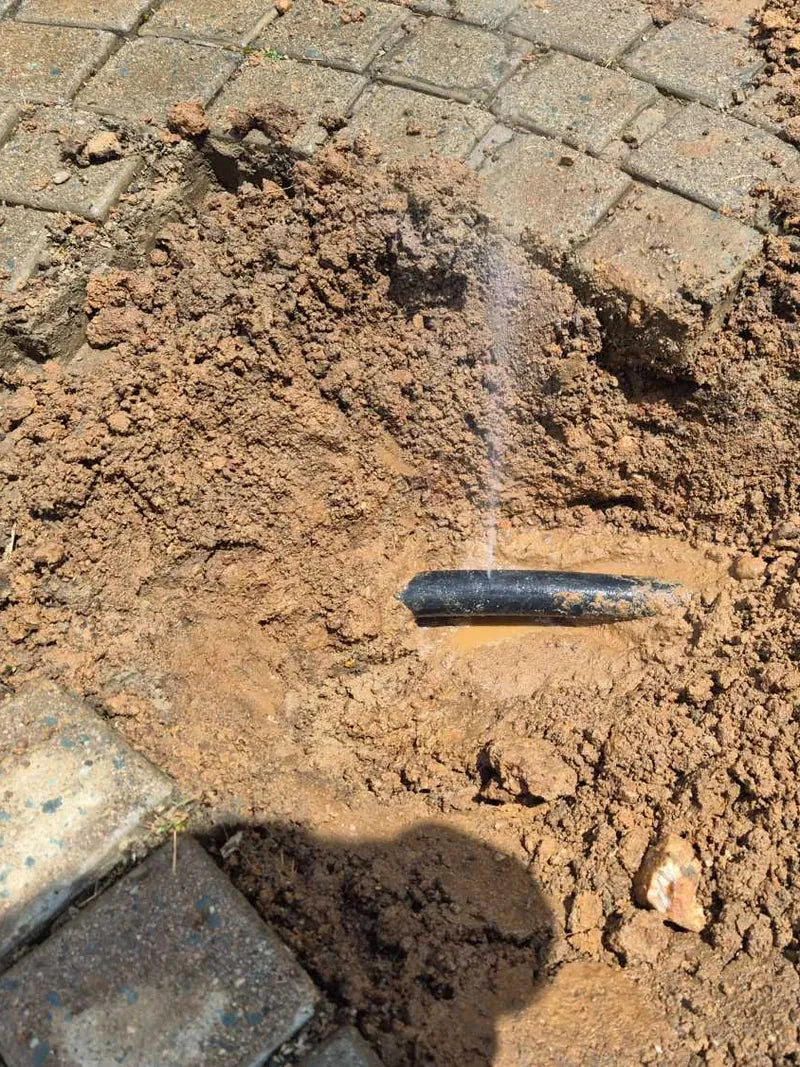 Water spraying from a damaged black pipe in a hole dug through paving