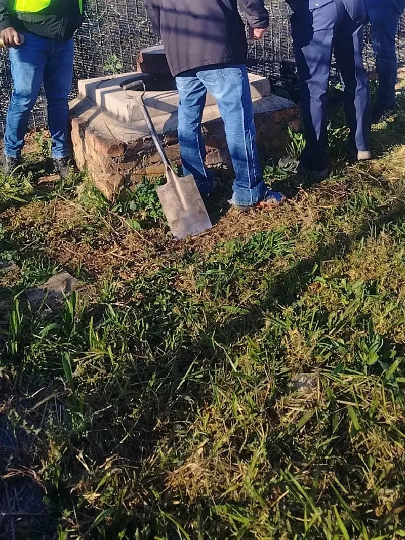 People in workwear standing around an open brick manhole on grass beside a security fence