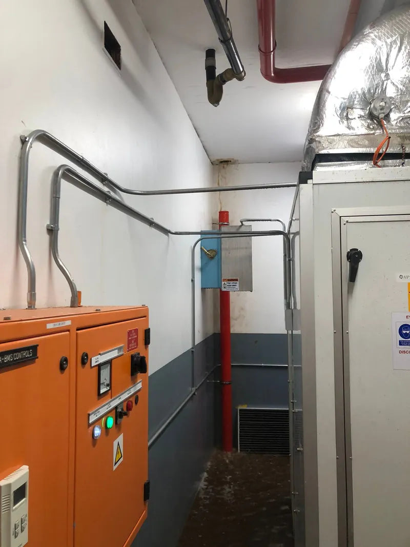 A generator plant room with an orange control panel, steel conduit and a generator enclosure