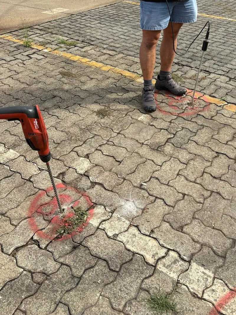 A technician with a tracer-gas probe and a drill on car-park paving marked with red paint