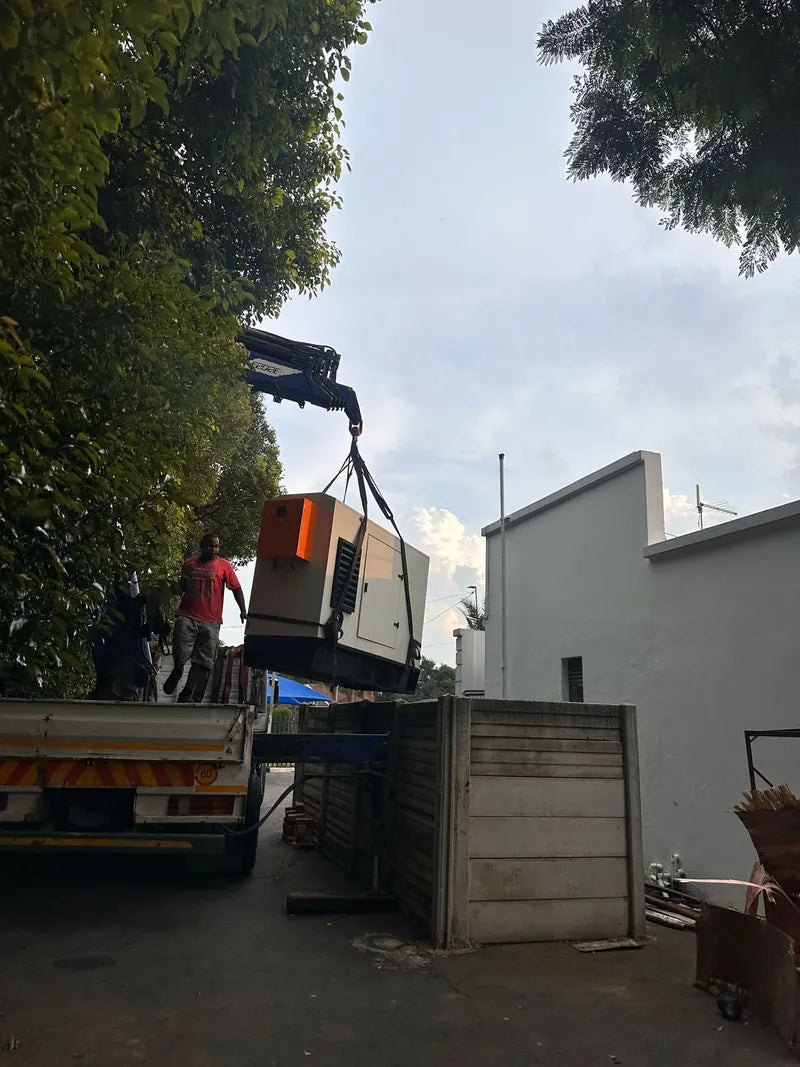 A crane truck lowering a large generator over a wall at a commercial property