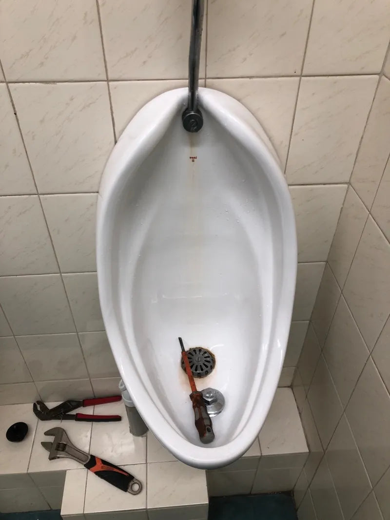 A wall-hung urinal with its waste fitting removed and plumbing tools beside it