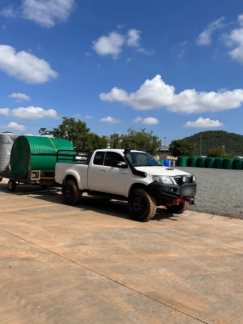 A white bakkie towing a green water tank trailer in an open yard