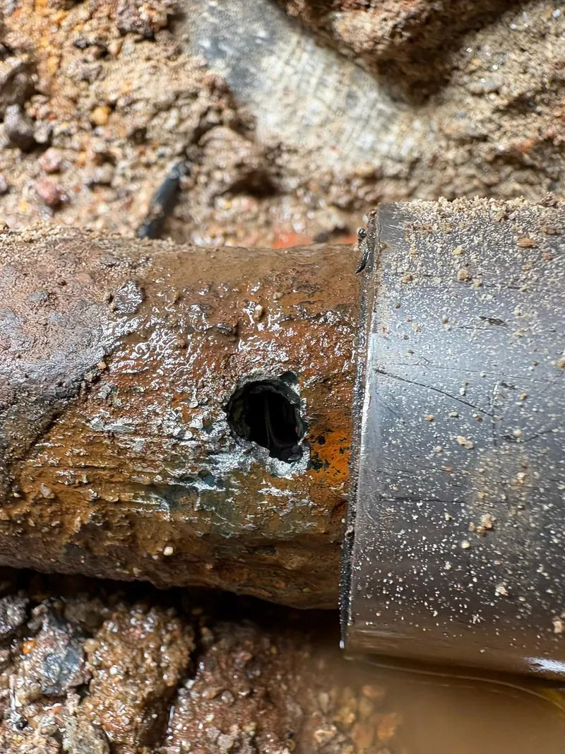 A corroded steel water pipe with a leak hole, exposed in an excavation