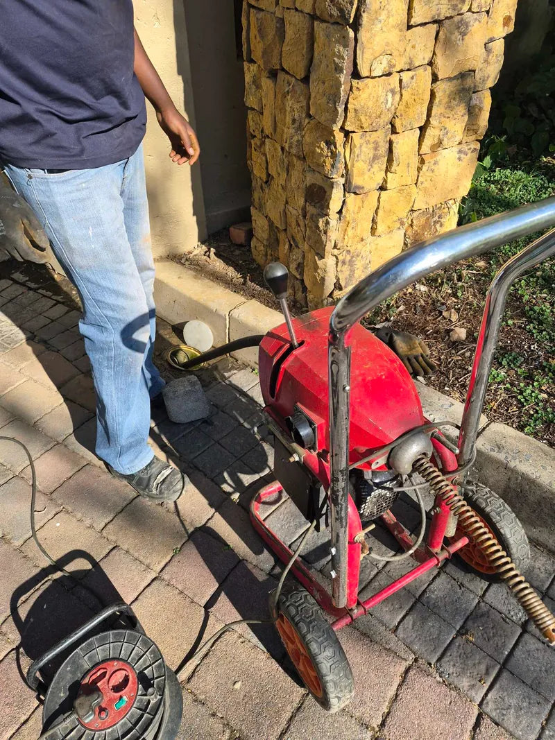 A technician standing beside a drain-cleaning machine with its spiral cable on brick paving