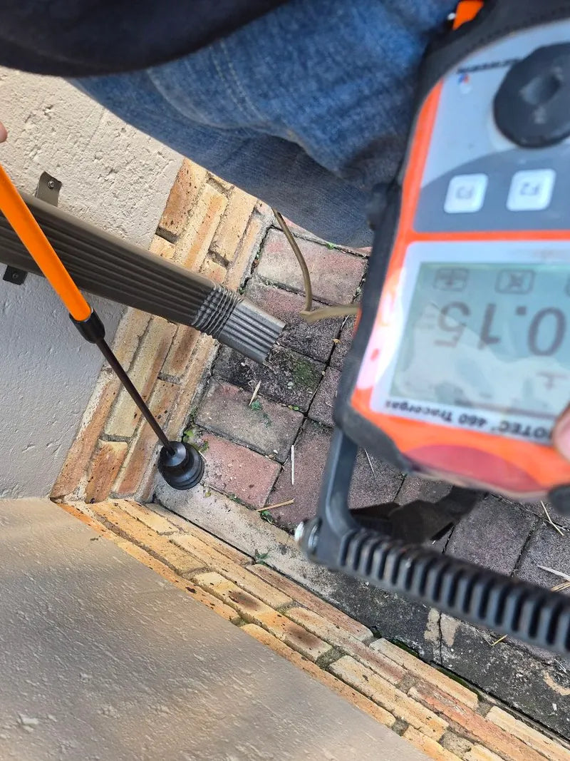 A tracer-gas leak detector and probe held over paving beside a wall