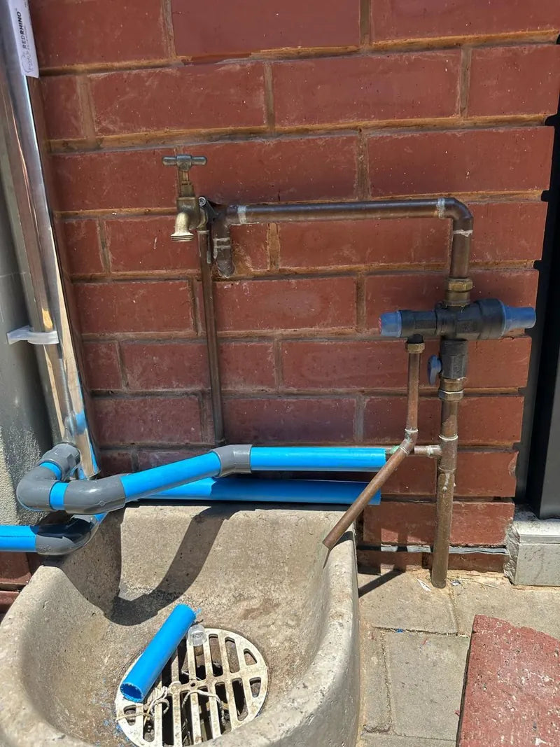 Blue water pipes, brass taps and a valve on a brick wall above a drain trough