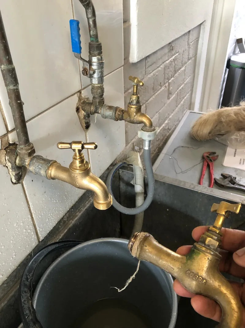 A hand holding a brass tap at a laundry basin below wall-mounted taps and pipes