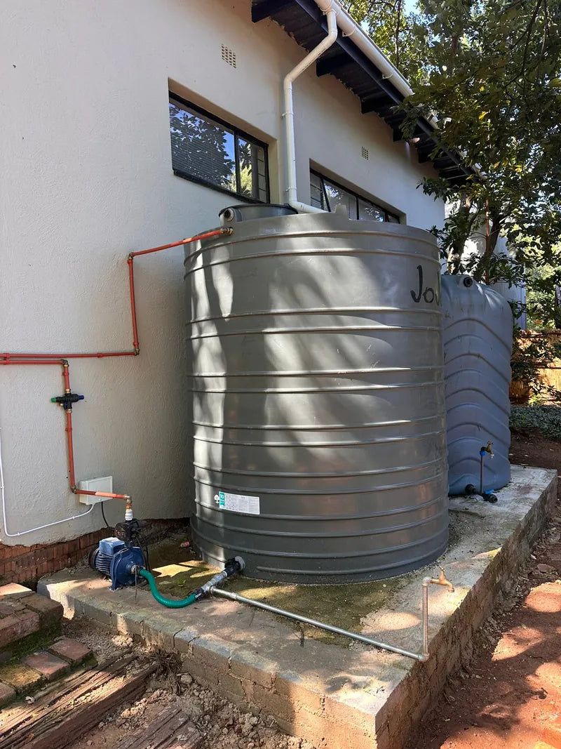 A grey JoJo water tank on a raised base beside a house, with a pump and pipework