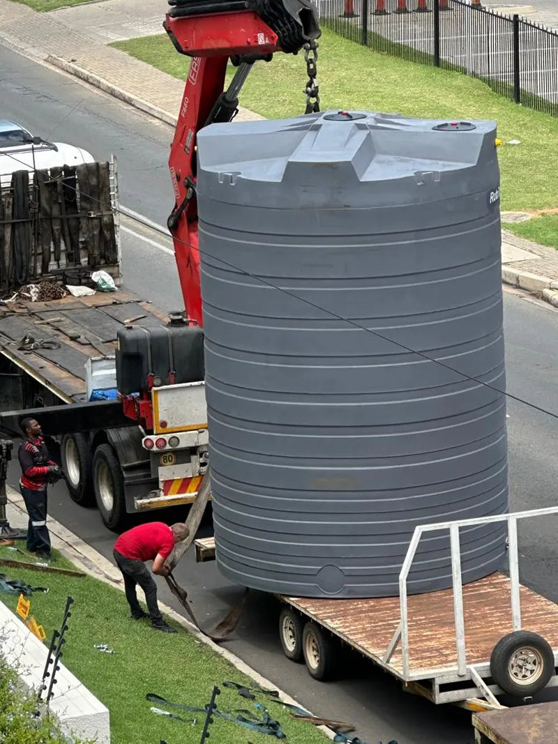A truck-mounted crane lifting a large grey water tank off a trailer at the roadside